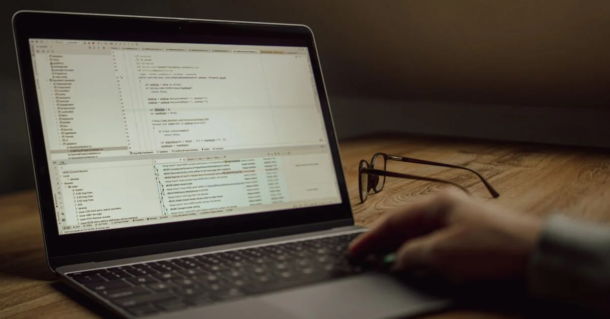 Une personne travaille sur du code informatique sur un ordinateur portable à l'intérieur. Des lunettes sont posées sur la table.